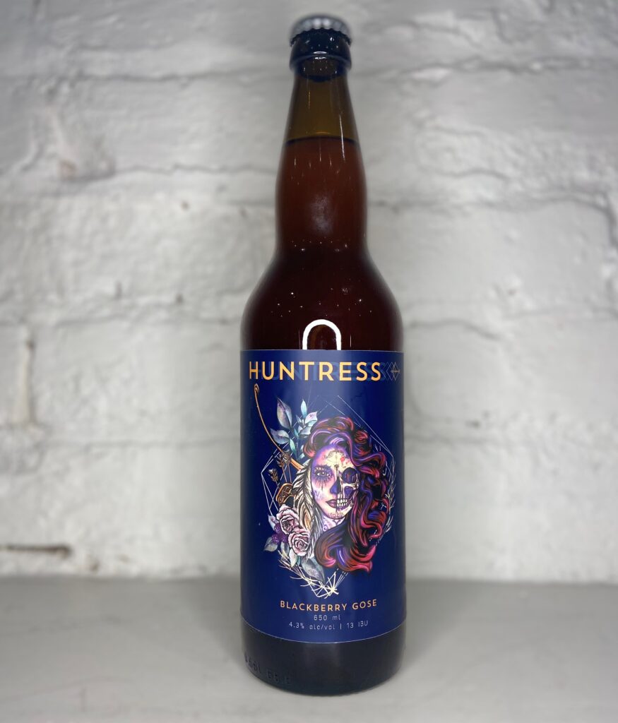 A bottle of 'Huntress,' a beer from Amsterdam Brewing featuring a dark blue label with a woman with half her face made up like a skull and long flowing pink and purple hair.  The style 'Blackberry Gose' is listed below the image.