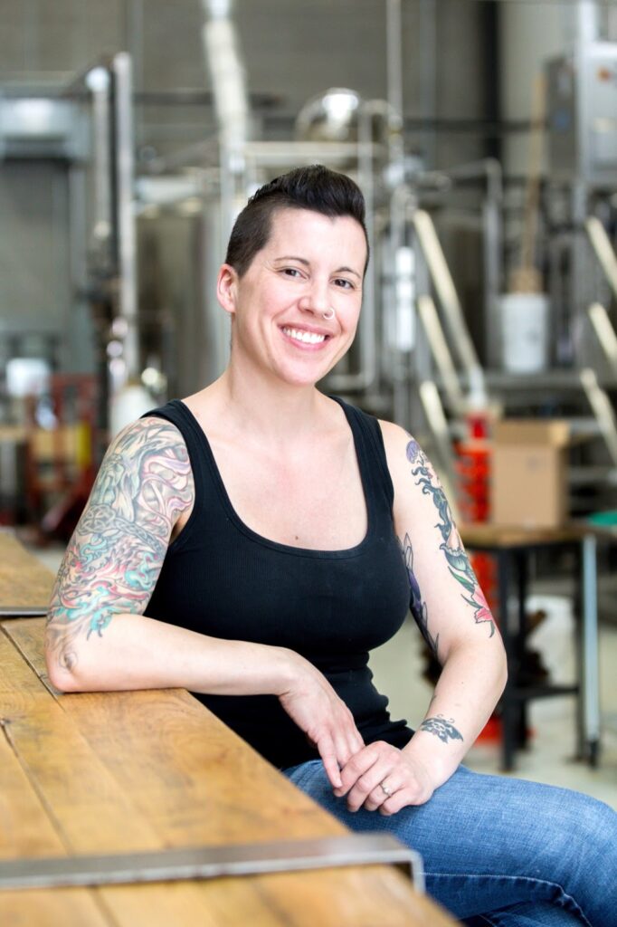 A photo of Erin sitting at the bar at Little Beast's Brewery smiling at the camera.  She has short dark hair, a nose ring and colourful tattoos on both arms and is wearing jeans and a black tank top. 