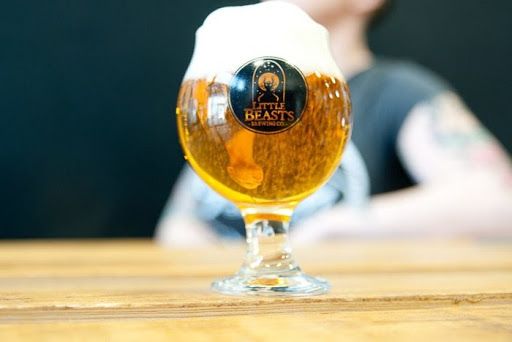A glass of beer sitting on a wooden bar with the Little Beasts logo on the glass.  Erin is out of focus in the background. 