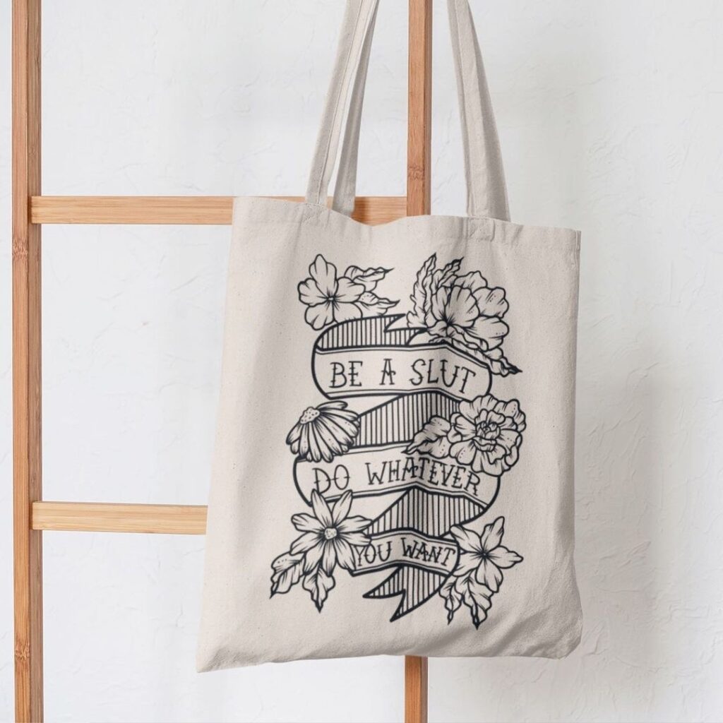 A photo of a tote back with Lucky Little Queer's artwork on it.  The tote is beige and the artwork is a black line drawing of a ribbon with lovely flowers around it that says 'Be a Slut, Do Whatever You Want' in all caps.
