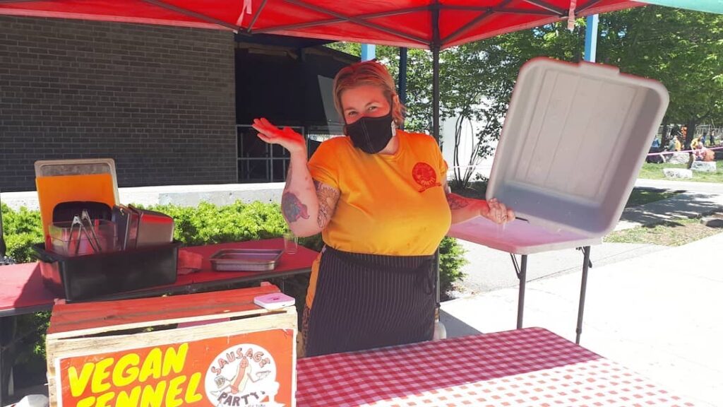 A photo of Emily (owner of Sausage Party) standing outside in her businesses booth.  She is wearing a bright yellow tshirt and a black mask and holding an empty bus bin, posing with 'ta dah!' arms. 